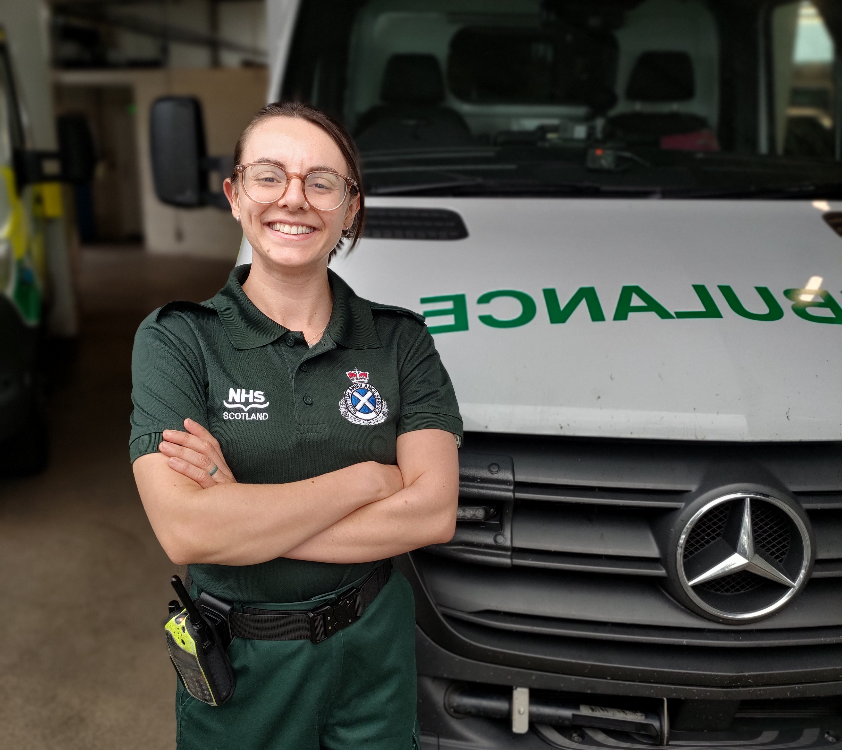 A female paramedic stood smiling with her arms crossed in front of a parked ambulance.