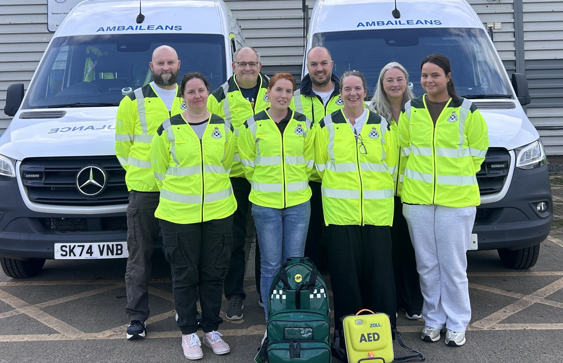 Eight community first responders dressed in yellow high vis stood in front of two ambulance vehicles.