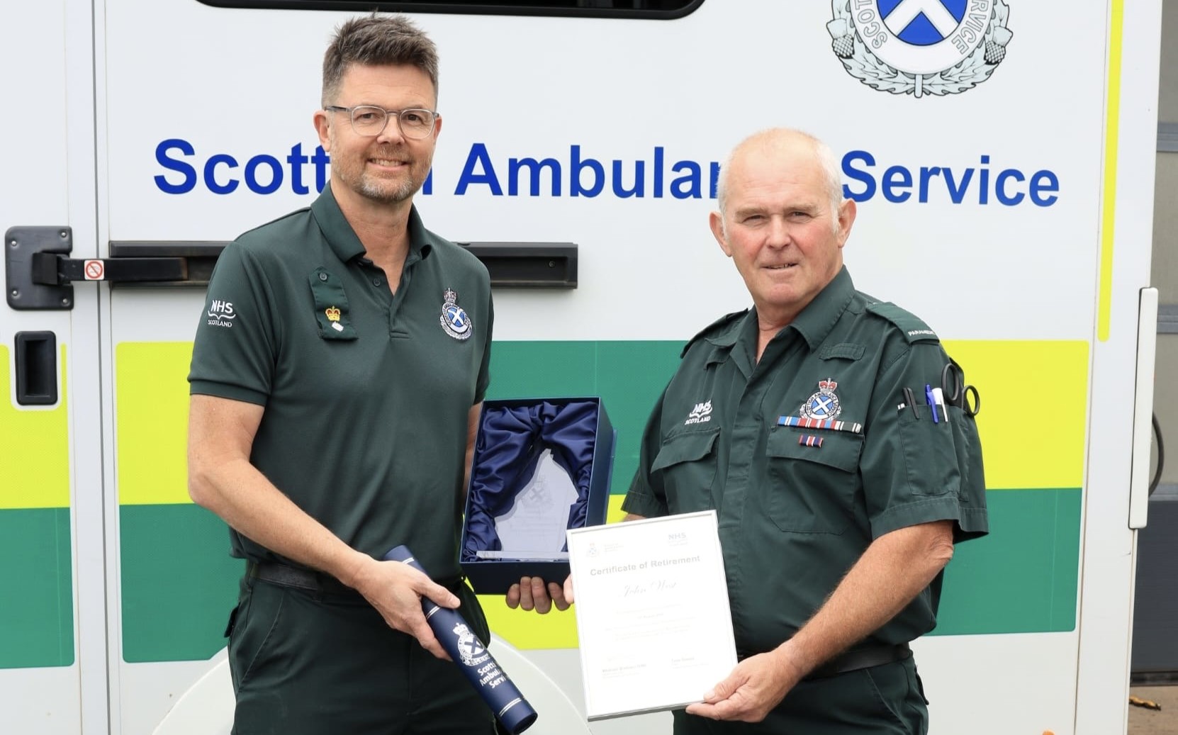 Two Scottish Ambulance Service staff members stand in front of a Scottish Ambulance Service vehicle. One person presents a retirement certificate and commemorative gift to a colleague wearing a green NHS Scotland ambulance uniform with service ribbons. The ambulance vehicle and service branding are visible in the background.