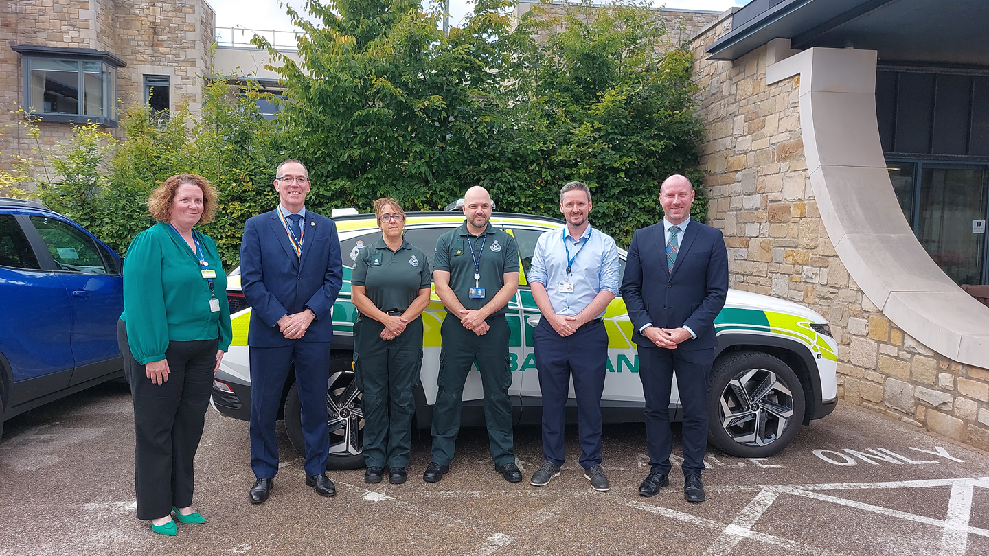 Group of 6 people standing in front of a parked ambulance car including SAS Chief Executive Michael Dickson and SG Cabinet Secretary Neil Gray.