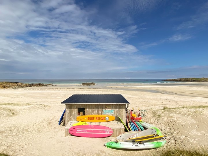 A beach shack on a beach with surf boards in front and the sea in the distance