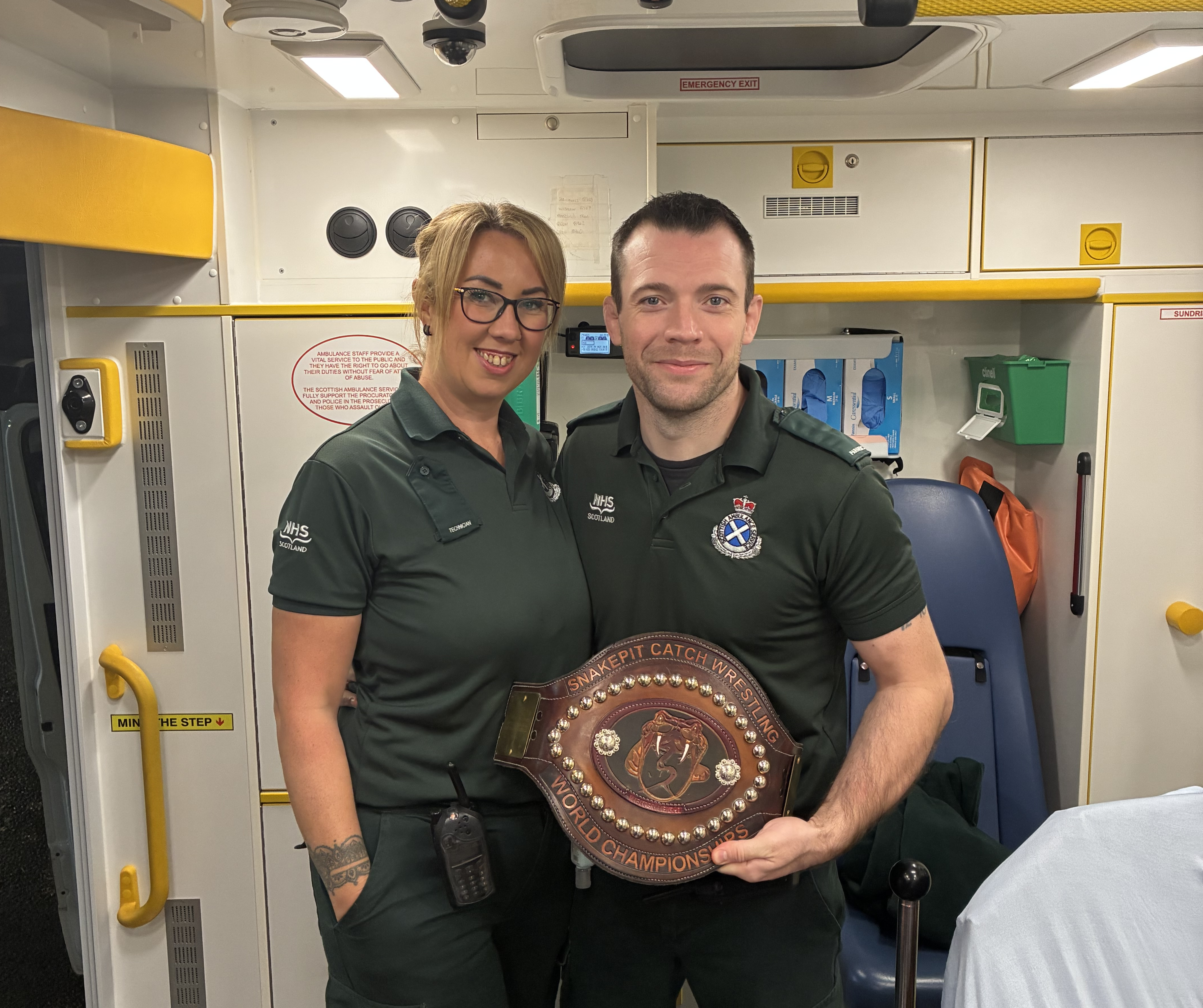 A female and male SAS crew member dressed in dark green uniforms stood in the back of an ambulance. The man is holding a Catch Wrestling World Championship belt.