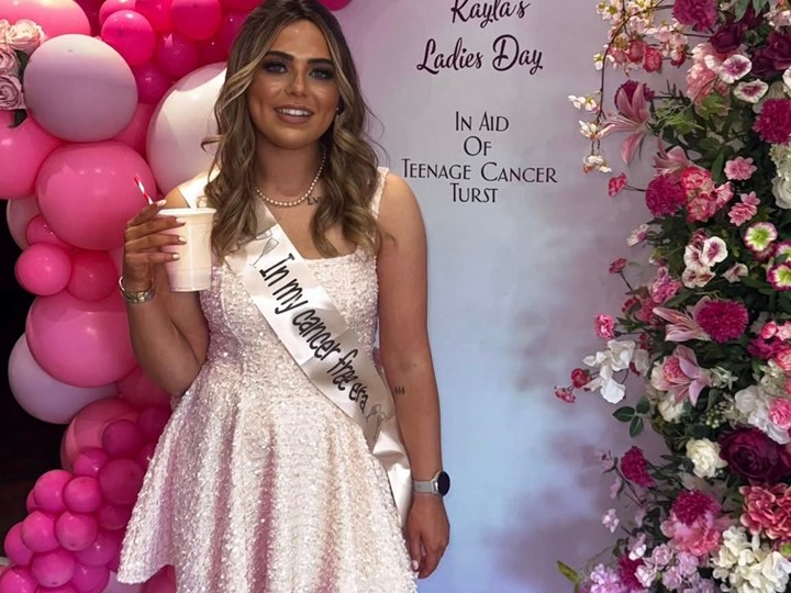 Kayla stands with a cup smiling in front of a fundraising board for the Ladies' Event