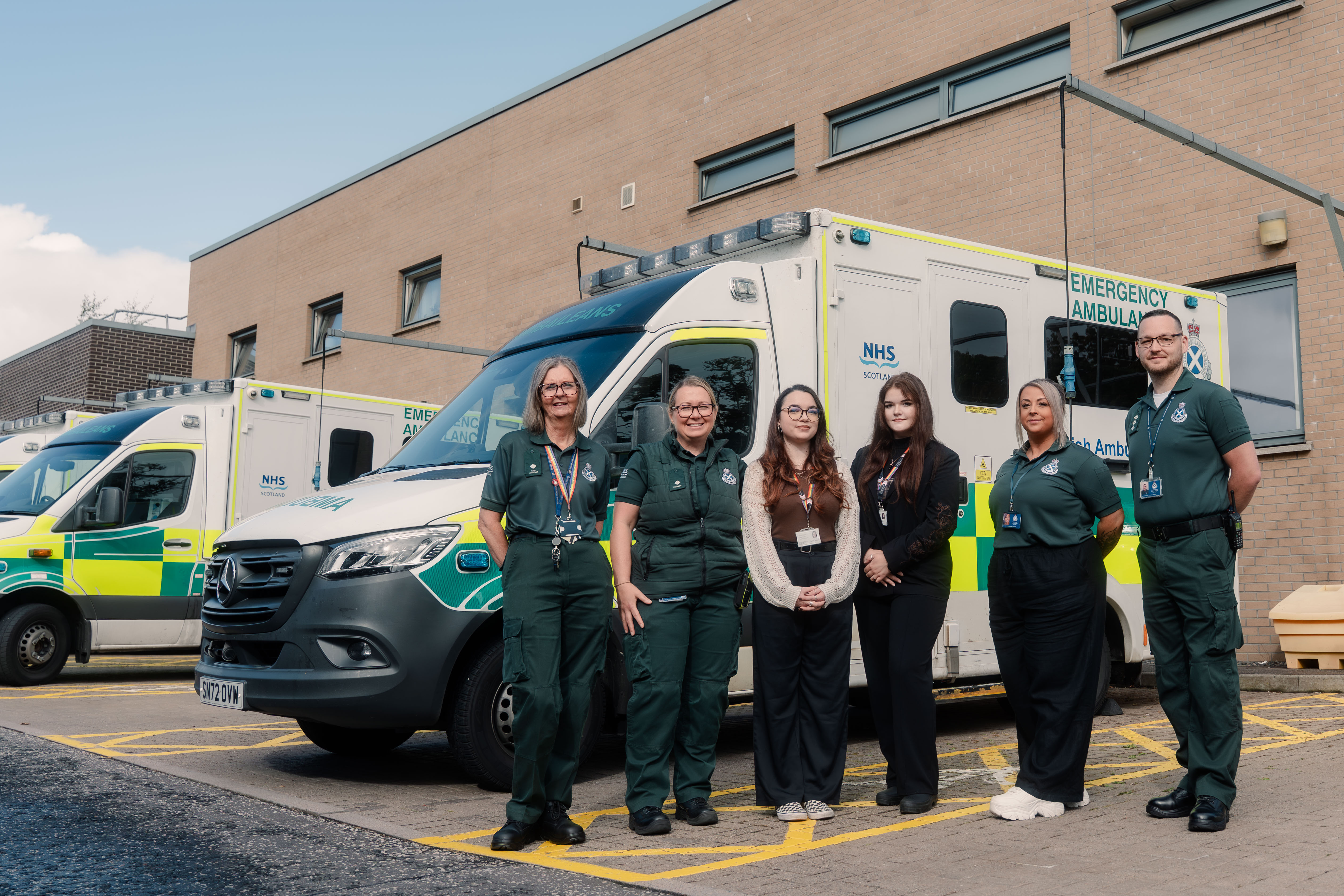 four paramedics and two people standing in front of a parked ambulance on a sunny day.