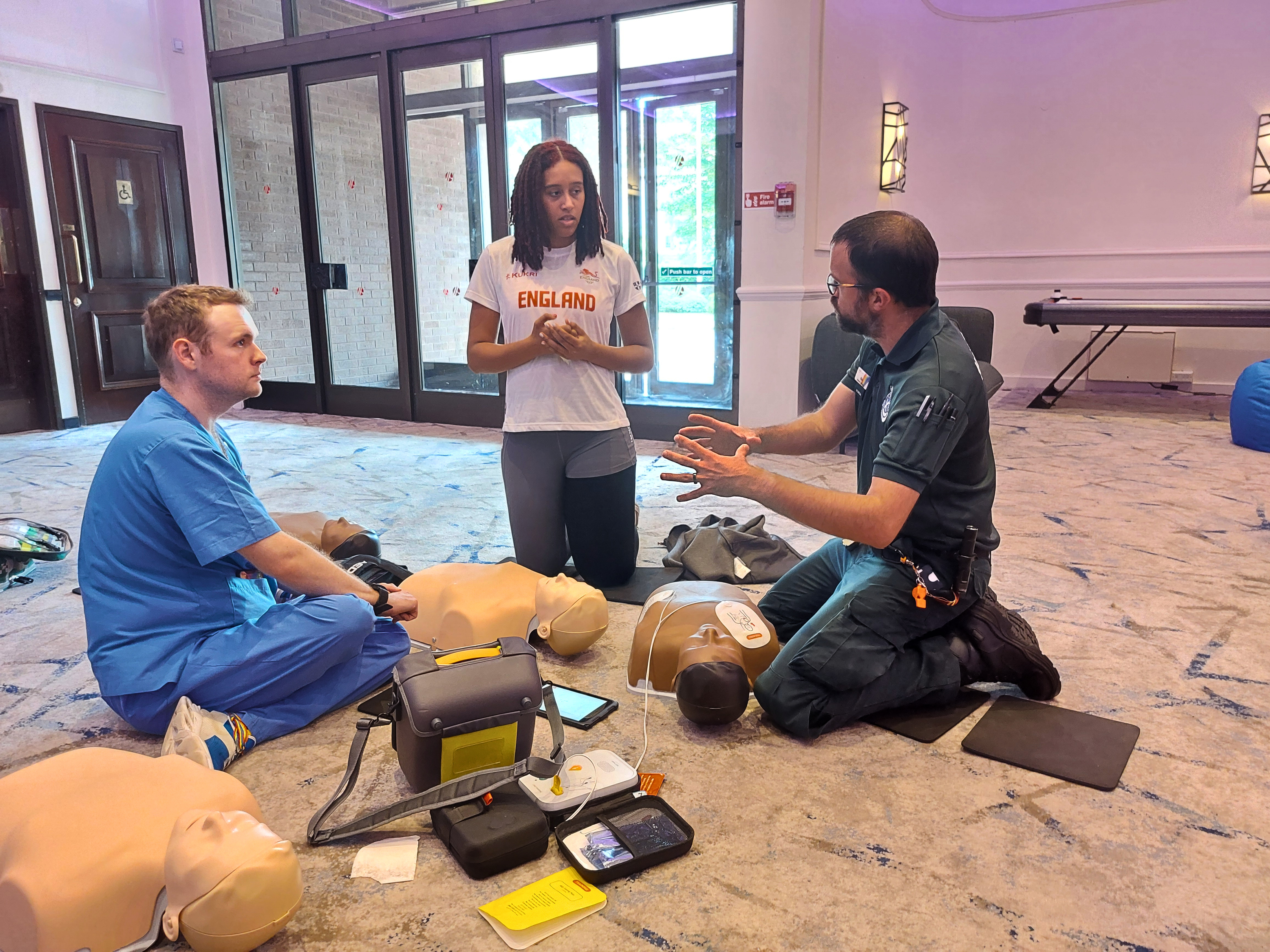 Two medical professionals instructing an athlete on how to perform CPR on a dummy.