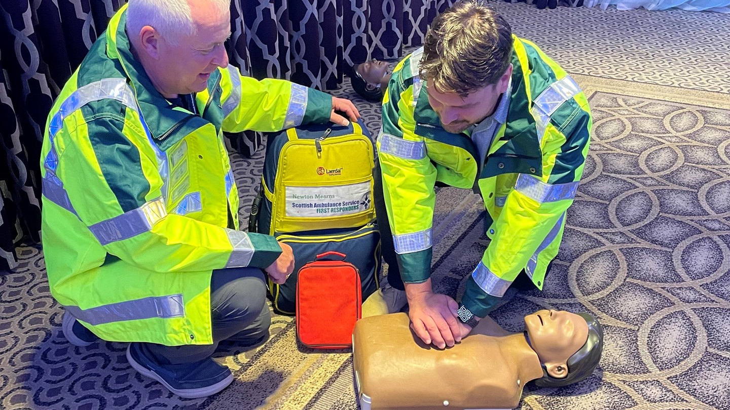 Two community first responders in high vis jackets demonstrate CPR on a CPR dummy,