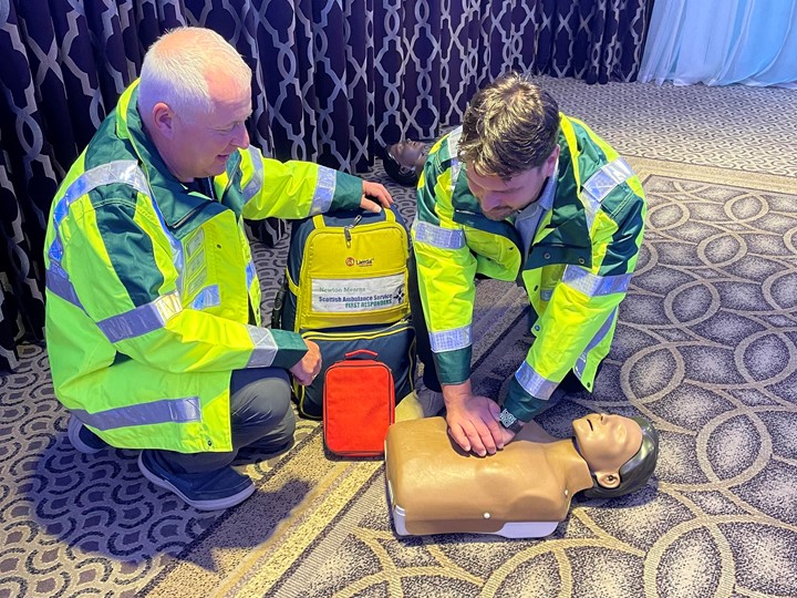 Two community first responders in high vis jackets demonstrate CPR on a CPR dummy,