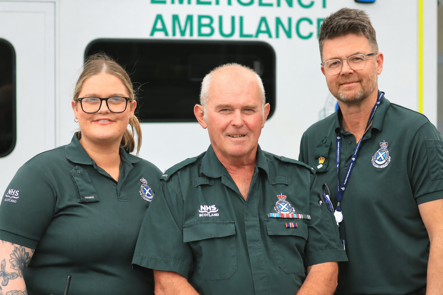 Three Scottish Ambulance Service staff members stand in front of an emergency ambulance. The person in the centre wears a green NHS Scotland ambulance uniform with service medals, while the two colleagues on either side wear green Scottish Ambulance Service polo shirts. The ambulance vehicle is visible in the background.
