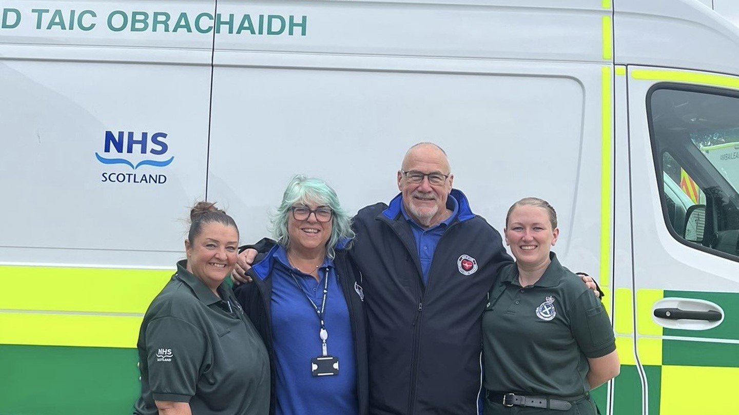 Sam Grieve, Mary and John Kinninmonth, and Laura Cameron standing together, smiling, in front of a parked ambulance vehicle