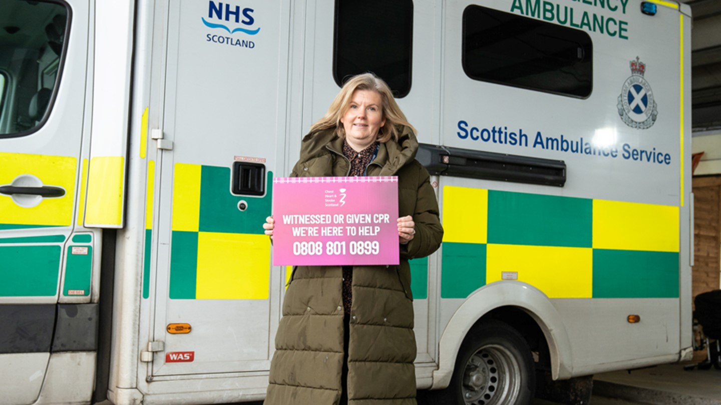 Lynsey Duncan holds a sign for CPR Bystander Support service whilst stood in front of an ambulance.