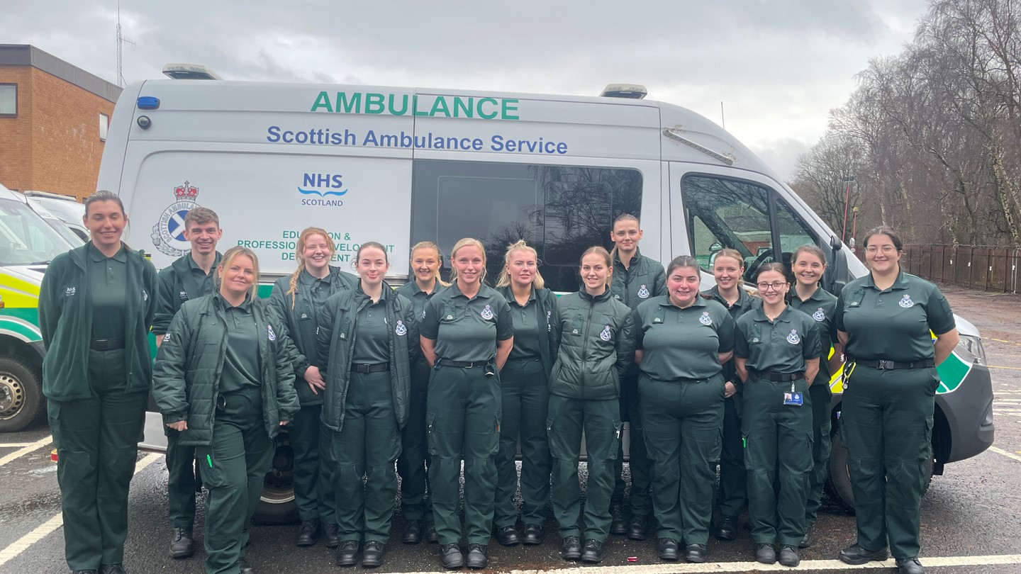 A large group of newly qualified paramedics in dark green SAS uniform stood in front of a parked ambulance van.