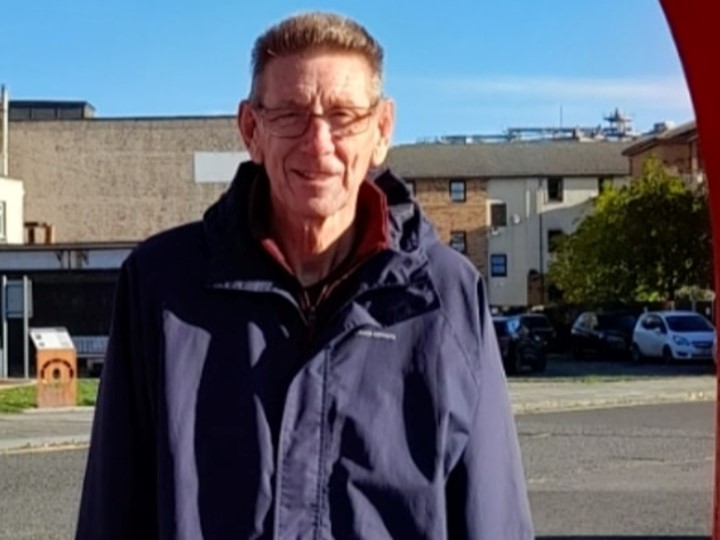 Man smiling at the camera on a sunny day behind him is a high street