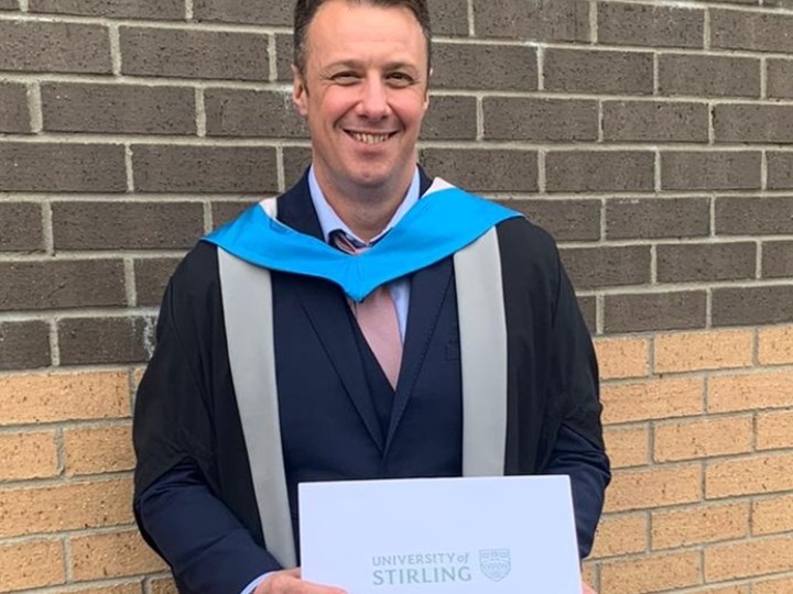 Dr Chris Aitchison wearing is graduation robes holds his certificate
