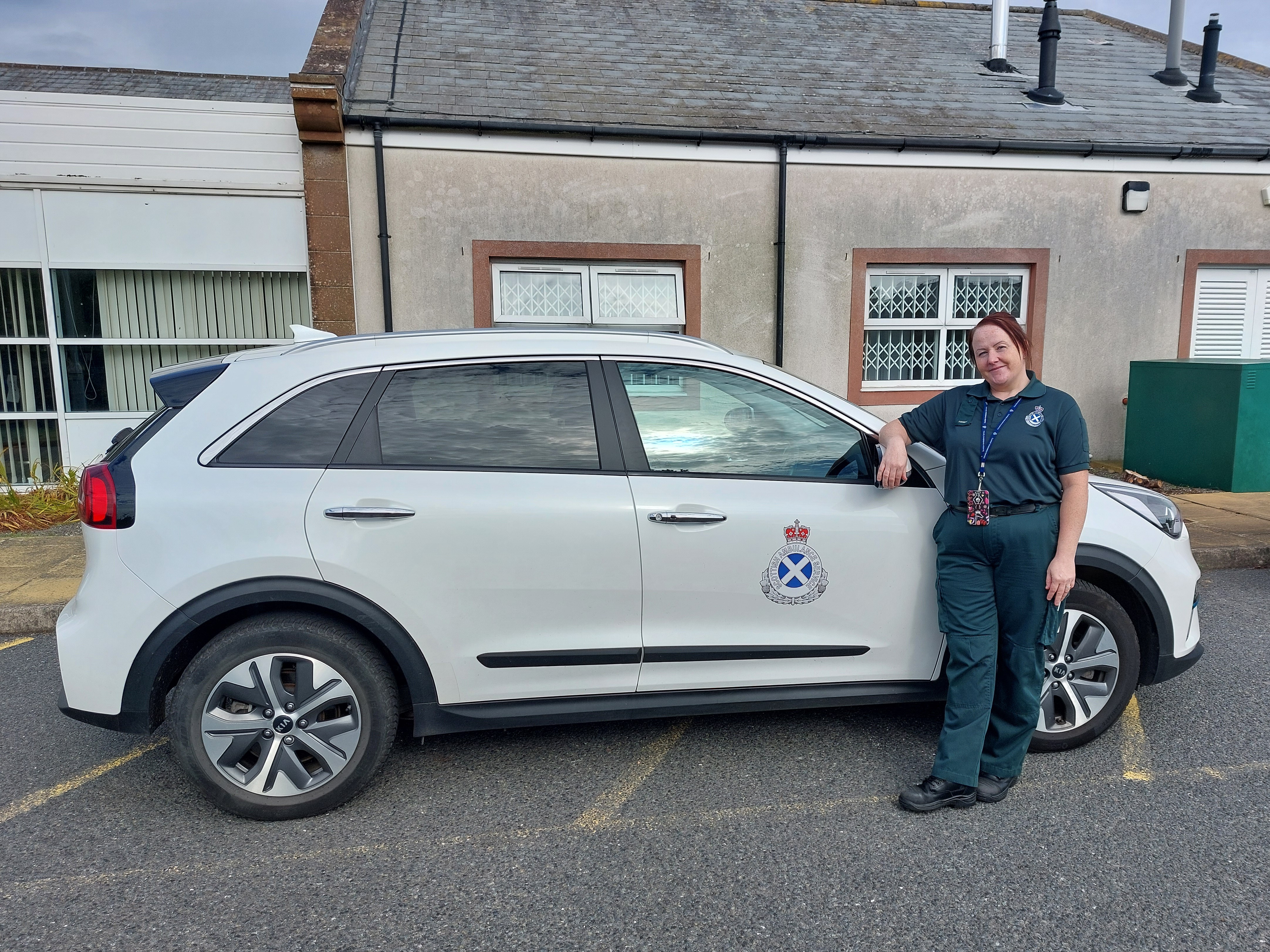 Paramedic in green uniform stood leaning on a white paramedic car that's parked in front a small building.