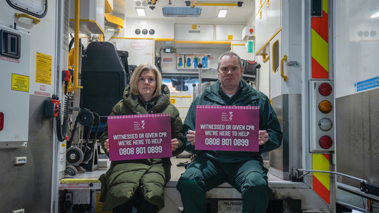 Lynsey Duncan and Steven Short hold signs for the CPR Bystander Support service whilst sat at the back of an ambulance with its doors open.
