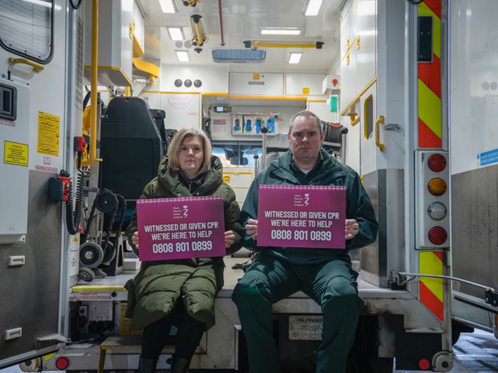Lynsey Duncan and Steven Short hold signs for the CPR Bystander Support service whilst sat at the back of an ambulance with its doors open.
