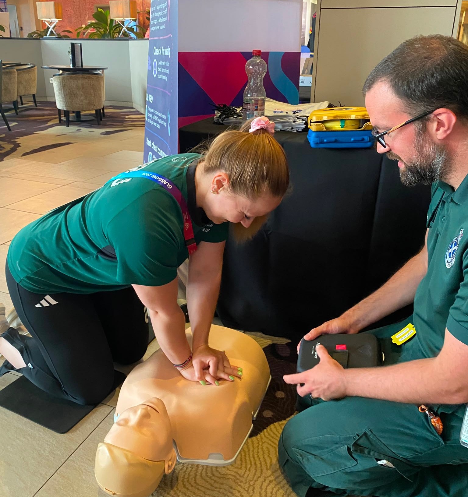 An athlete in green performing CPR on a training dummy while a paramedic looks on.