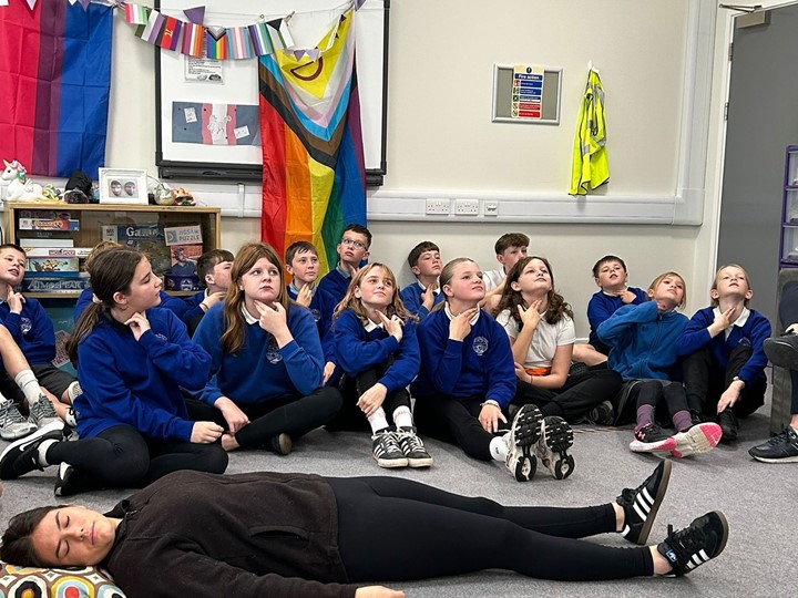 A woman laying on the floor pretending to be asleep in front of a full class of P7 pupils who are holding their hands to their throats to count their pulse.