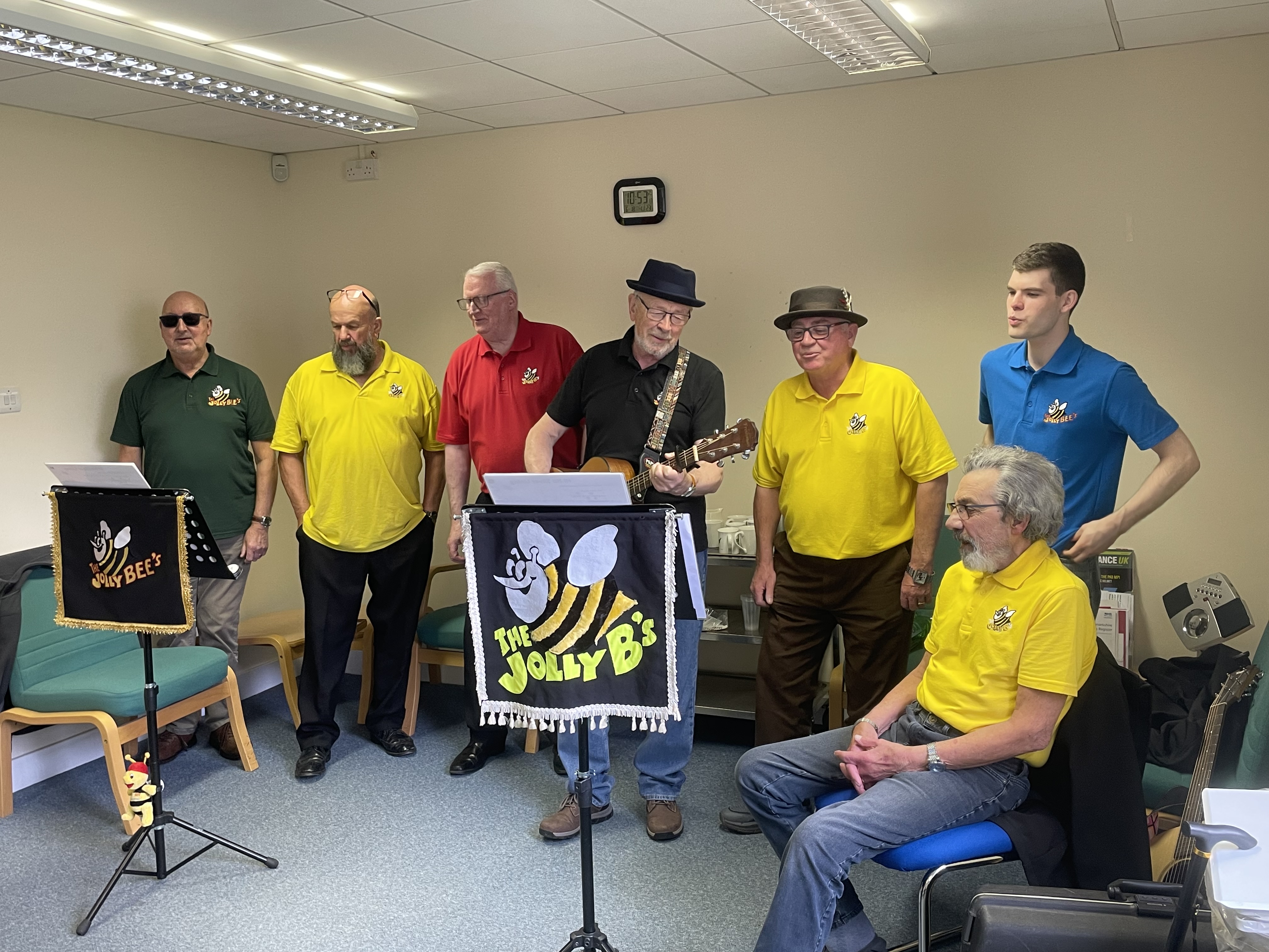Seven men in brightly coloured polo t-shirts singing in front of two banners with a bee and text saying The Jolly B's.