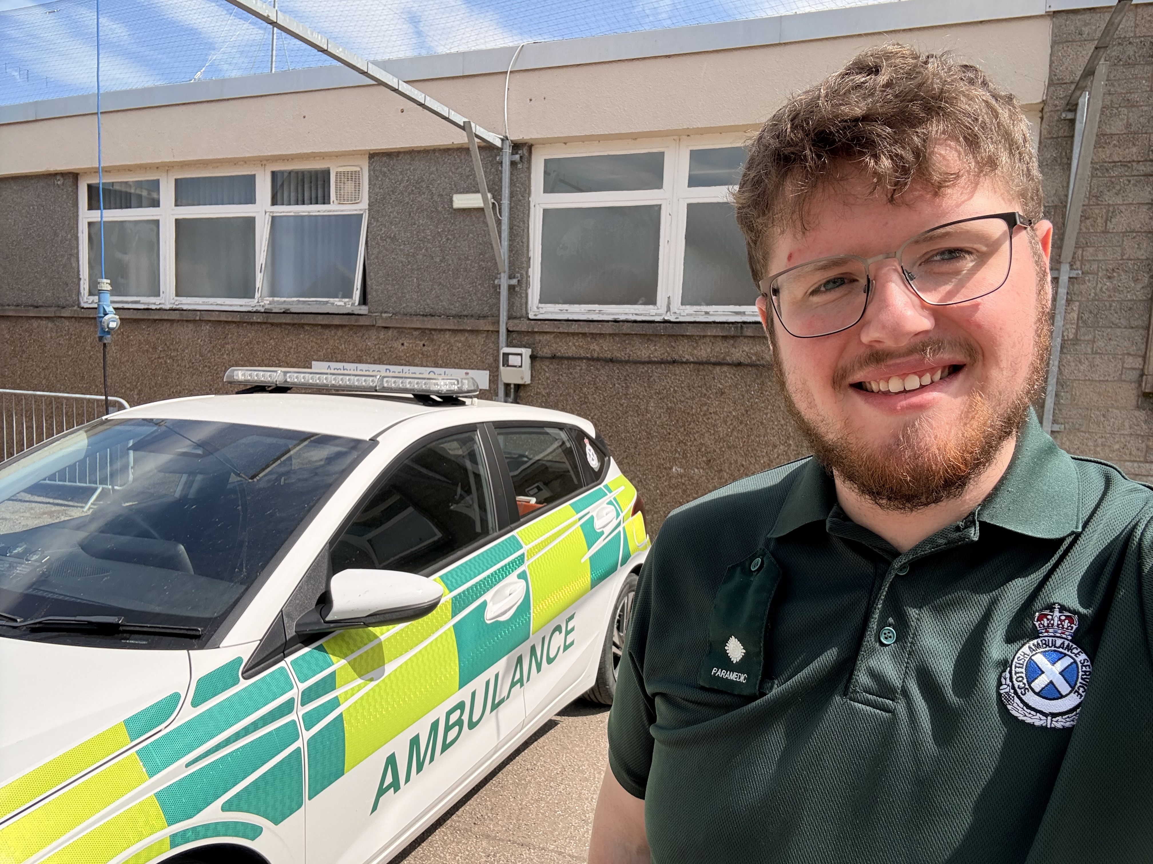 Paramedic in green uniform taking a selfie next to a parked ambulance outside a building on a sunny day.
