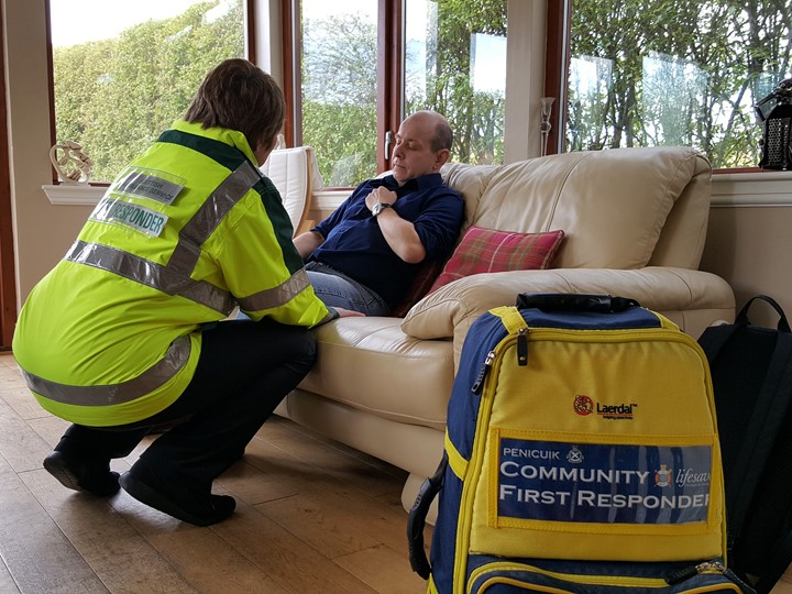 Female community first responder kneels while treating a male patient who is sat on a sofa.