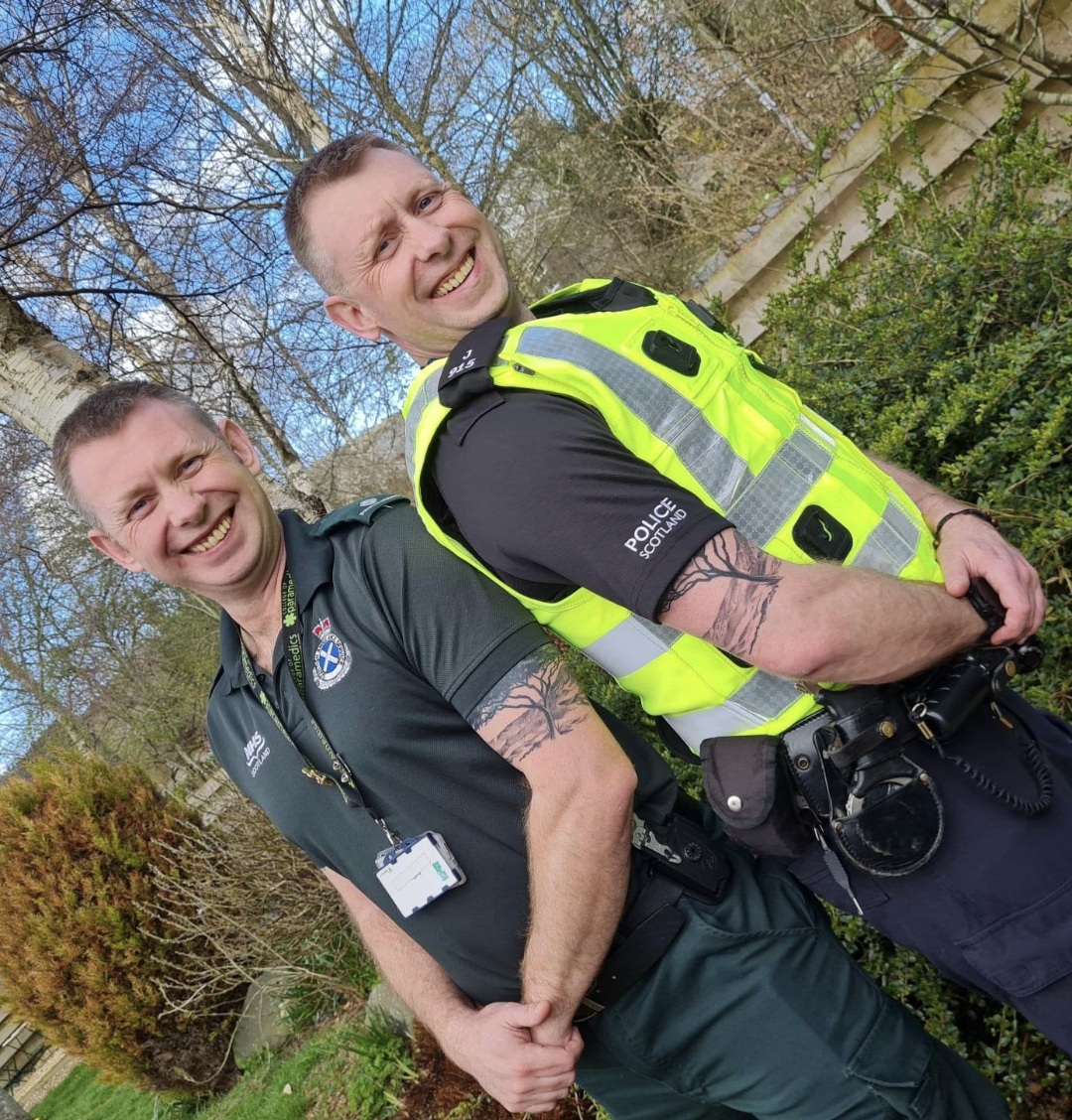 Two twin brothers smiling and stood back to back. One dressed in Ambulance uniform and the other dressed in Police uniform.