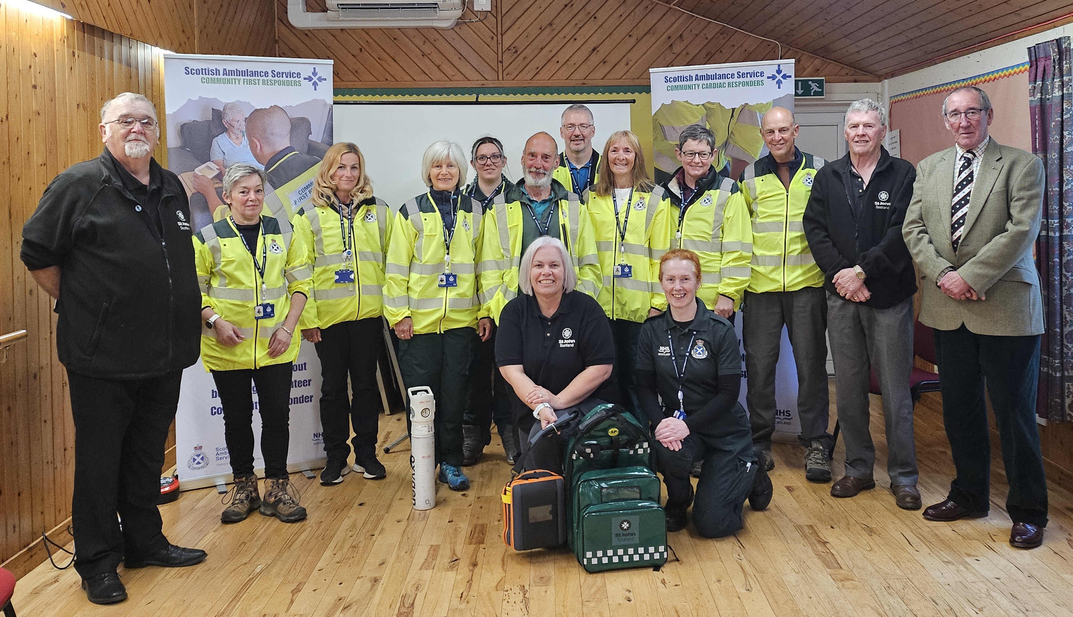 Group of emergency responders and volunteers in high-visibility jackets posing indoors with medical equipment on the floor.