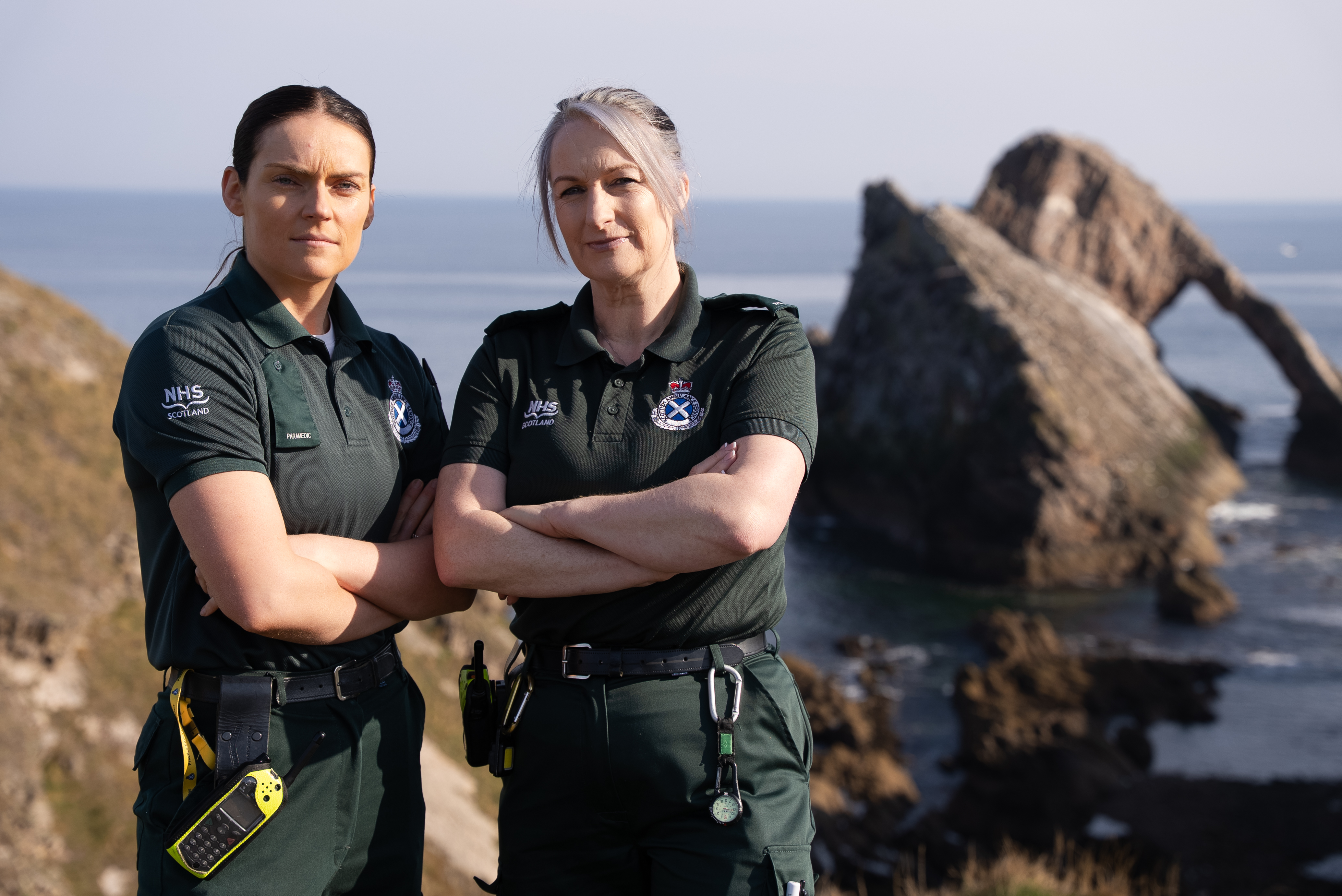 Two paramedics stand with arms crossed with dramatic sea background in the distance