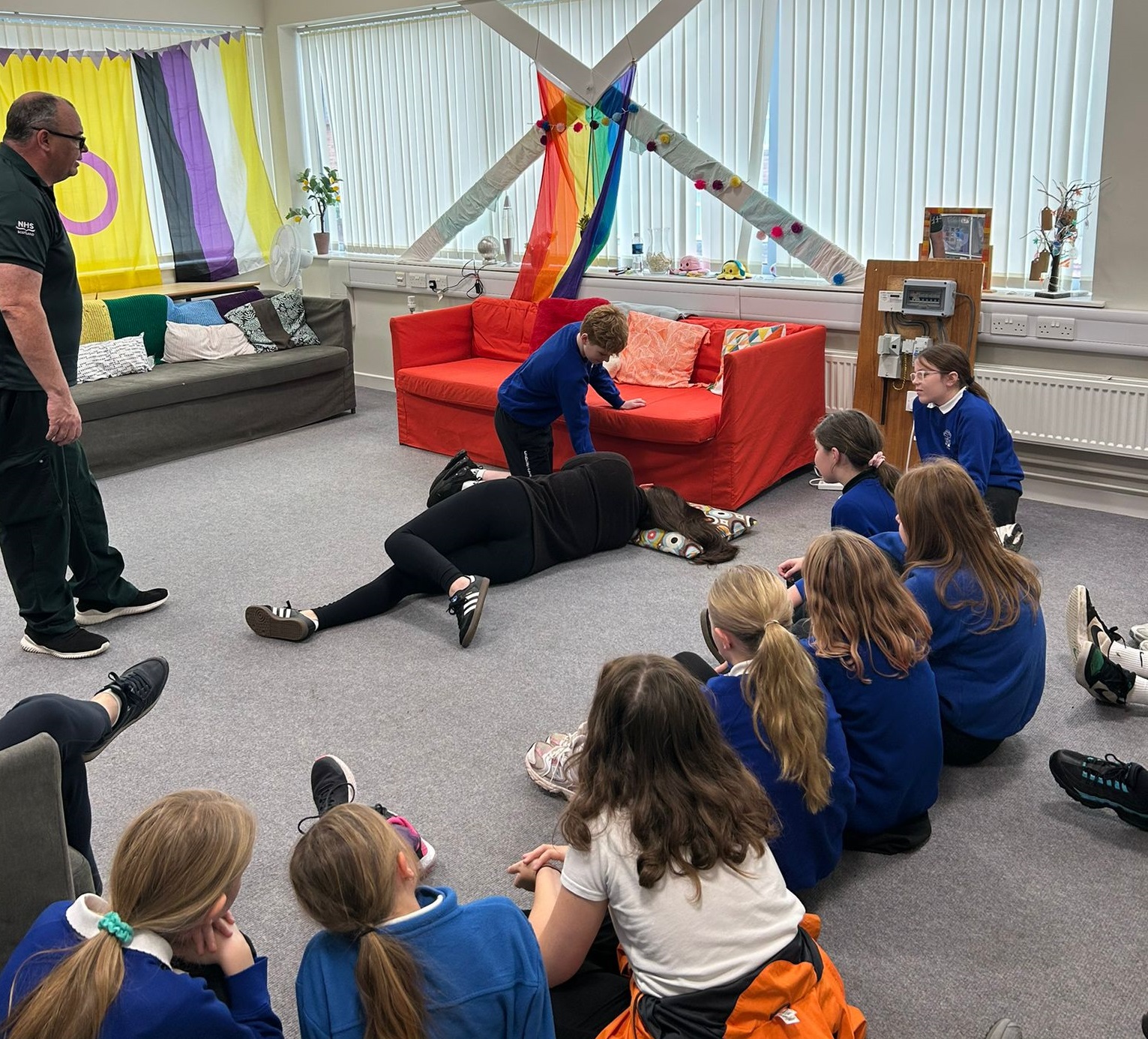A group of P7 pupils watching a demonstration of how to put a person into the recovery position whilst a SAS technician gives instructions.