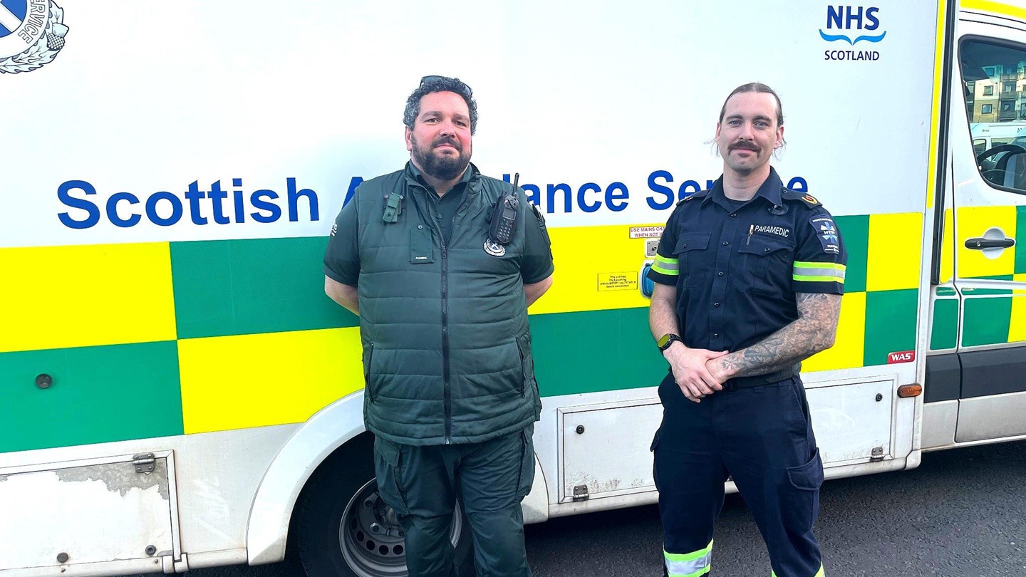 Paramedic Steve stands next to overseas staff member Luke in front of a Scottish Ambulance Service vehicle smiling