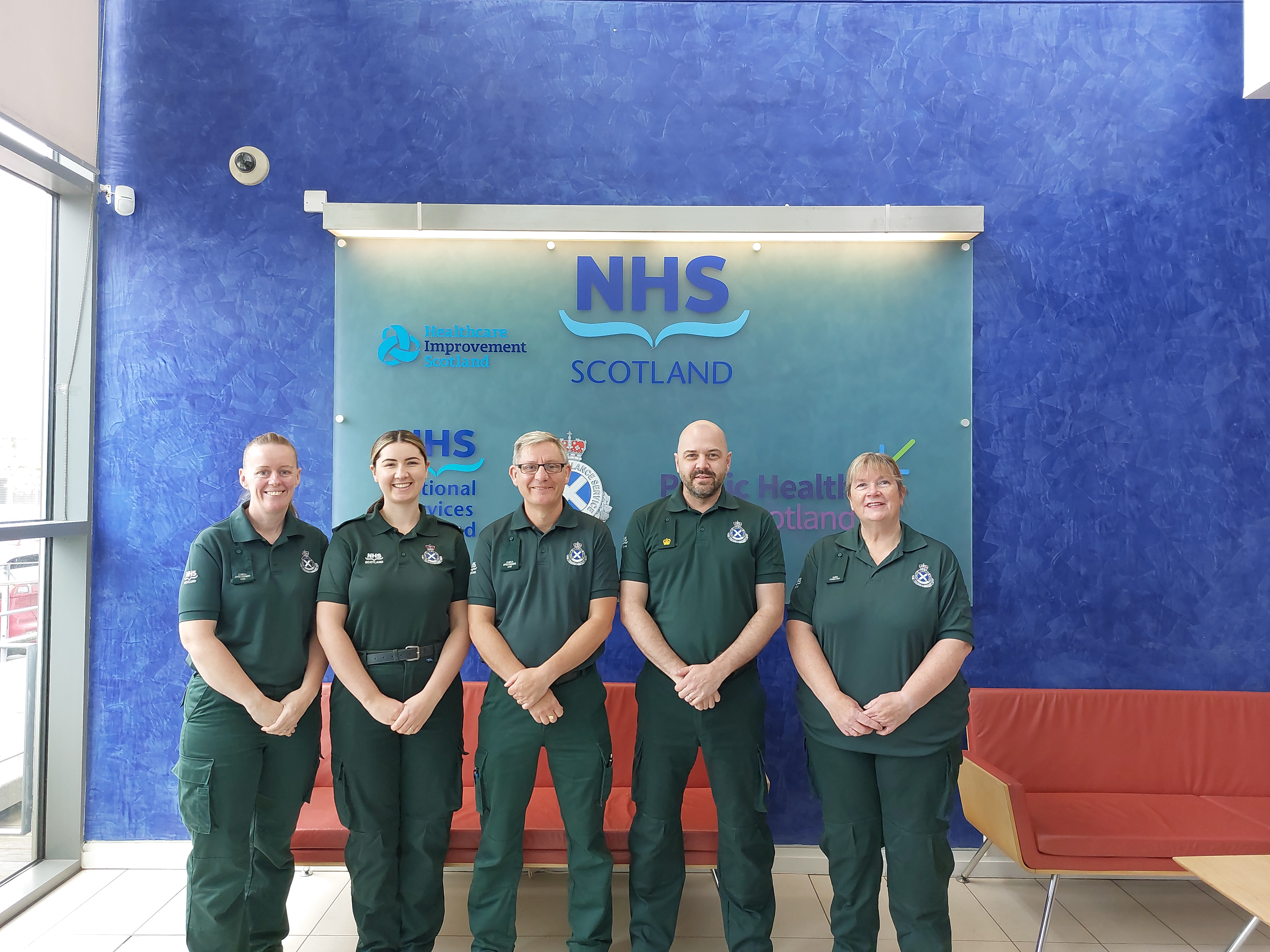 Five members of Ambulance staff in uniform stand in front of a Board with NHS Logos