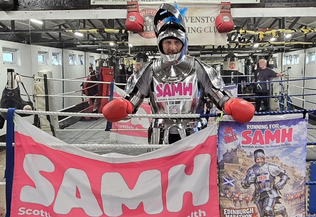 Person in a shiny silver knight armour with red boxing gloves holding a SAMH banner inside a boxing ring.