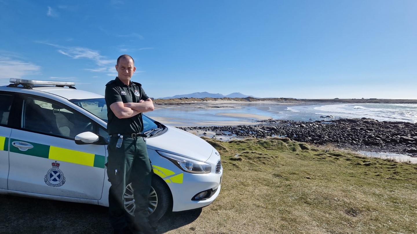 SAS Ambulance worker in uniform stood by a parked ambulance vehicle overlooking a beach