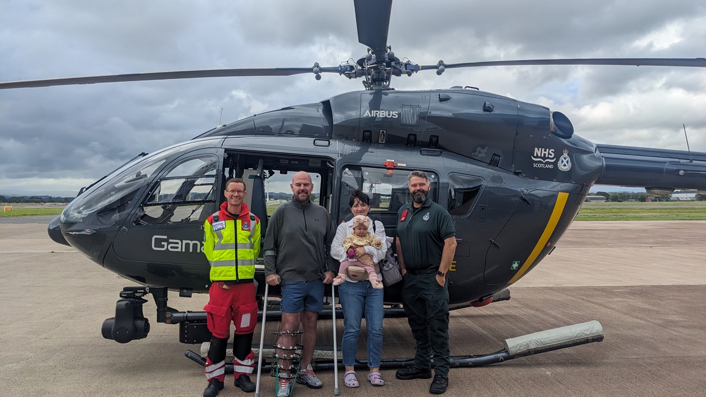 Four people and a baby stood together in front of a parked air ambulance helicopter on the tarmac at Glasgow Airport.