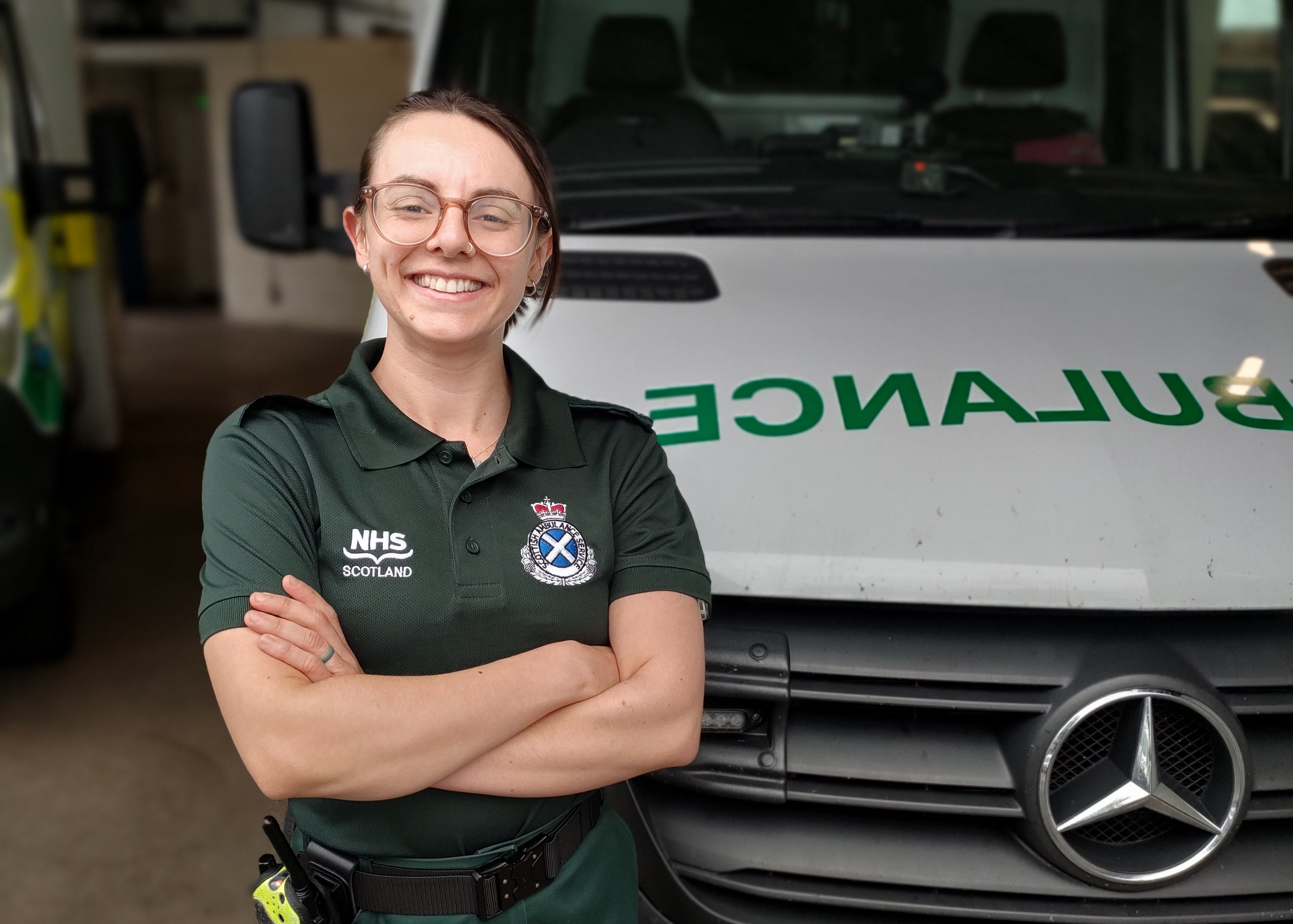 A female paramedic stood smiling with her arms crossed in front of a parked ambulance.